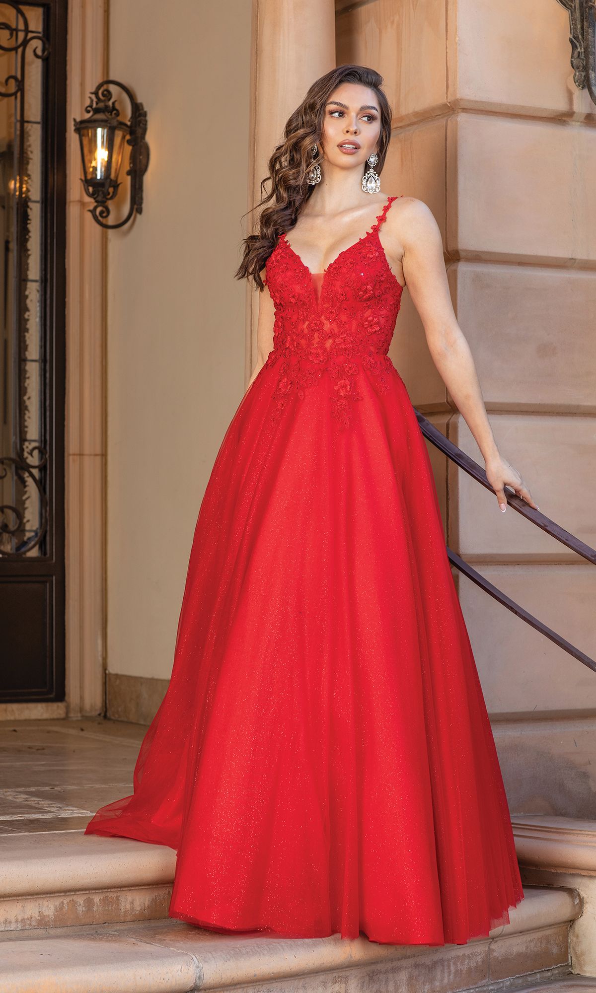 A woman in the Long Glitter Prom Dress: Dancing Queen 4328 stands on steps by a beige stone building, holding the railing, with wavy hair and statement earrings.